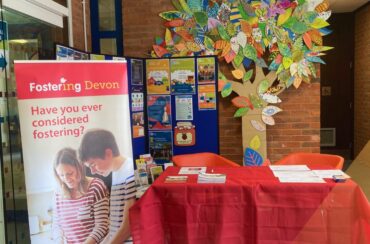 Picture of Fostering Devon set up at Barnstaple library. A red table with leaflets sits in front of a Fostering Devon pull up banner