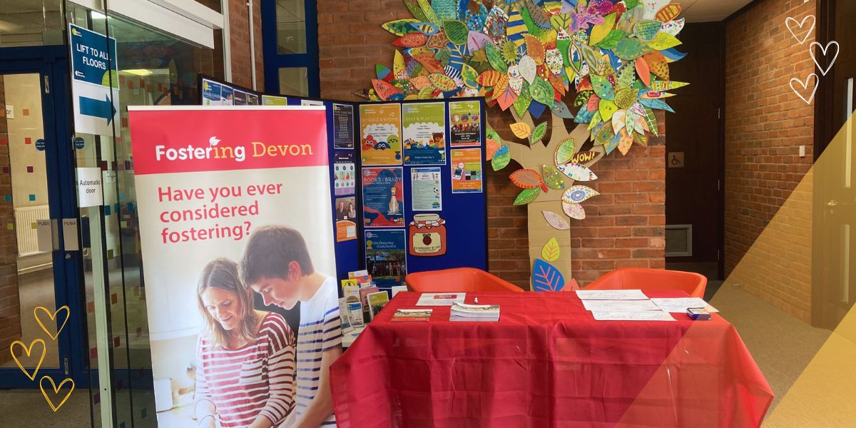 Picture of Fostering Devon set up at Barnstaple library. A red table with leaflets sits in front of a Fostering Devon pull up banner