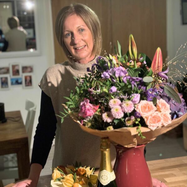 Sarah smiling at the camera, posing next to a bunch of flowers, a cake decorated with fruit, and a champagne bottle 