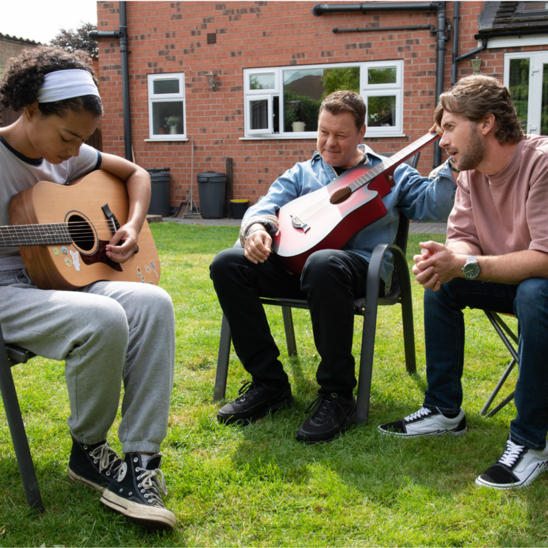 foster dad sat in the garden with foster daughter and foster son. The dad and the daughter are both playing on guitars. 
