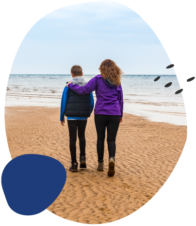Two children walk arm in arm along a sandy beach, seen from behind, with the sea and cloudy sky ahead.